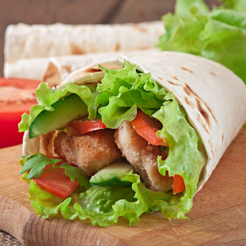 A John's Famous Bagels & Deli chicken wrap with lettuce, tomato, and cucumber on a wooden board.